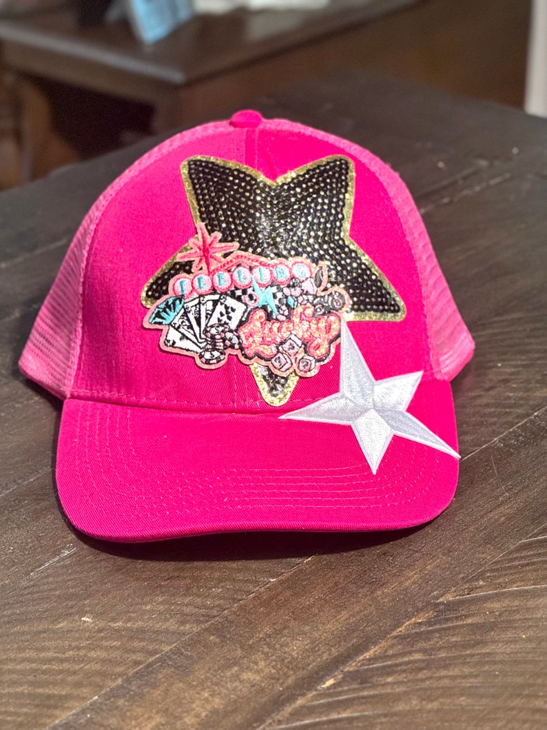 May include: A hot pink trucker hat with a mesh back. The front features a large star-shaped patch with black and gold embellishments, a white star, and a colorful graphic with the word "Lucky".