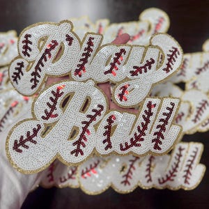 May include: A close-up of multiple white and gold sequined patches with the words "Play Ball" in red and white stitching. The patches are designed to look like a baseball.