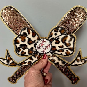 Può includere: Un fiocco decorativo a tema baseball con due mazze incrociate, ciascuna con una parte superiore marrone con paillettes e finiture dorate. Il fiocco presenta un motivo leopardato e una palla da baseball al centro con cuciture rosse, il tutto impreziosito da glitter dorati.