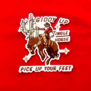Puede incluir: Parche bordado con un vaquero montando un caballo salvaje, un cactus y las palabras "Giddy Up", "Tingle Horse" y "Pick Up Your Feet". El parche tiene un borde blanco sobre un fondo rojo.