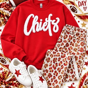 May include: A red crewneck sweatshirt with white fuzzy chenille lettering that reads Chiefs, paired with white jeans featuring a red and gold floral leopard print pattern and white sneakers with a red glitter star on the side of each shoe.