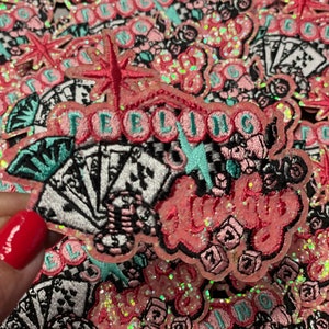 May include: Embroidered patches with the words "Feeling Lucky" in pink, with playing cards, dice, and starburst designs. The patches have a glittery, iridescent finish and are set against a background of similar patches.