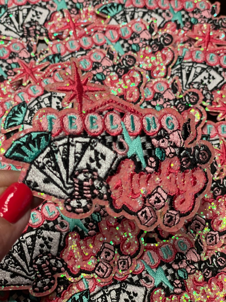 May include: Embroidered patches with the words "Feeling Lucky" in pink, with playing cards, dice, and starburst designs. The patches have a glittery, iridescent finish and are set against a background of similar patches.