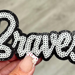 May include: A black patch featuring the word Braves in white script lettering. The letters are outlined with white sequins, and the background of the patch has a black glitter finish, creating a high-contrast decorative accessory for clothing.