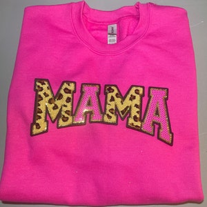 Może przedstawiać: Różowa bluza z dużym nadrukiem "MAMA" w różowych i złotych literach z wzorem lamparta.