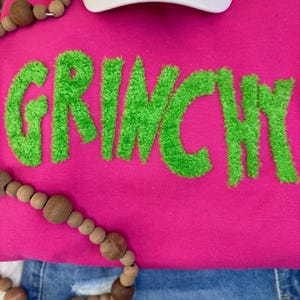 May include: A hot pink sweatshirt with the word "GRINCHY" in bright green, textured lettering. A white baseball cap and a wooden bead necklace are also visible. The sweatshirt is paired with blue denim jeans.