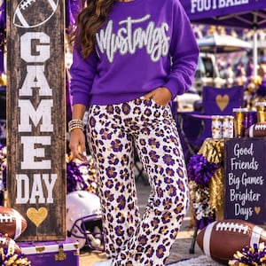 May include: A purple long-sleeve sweatshirt with the word Mustangs in silver script across the chest, paired with white trousers featuring a purple and gold leopard print pattern and white trainers with a purple star detail on the side.