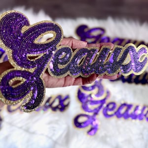 Puede incluir: Guion "Jeaux" con lentejuelas moradas y ribete dorado. Las letras están adornadas con pequeñas lentejuelas brillantes. El diseño se sostiene en una mano, con otros diseños similares visibles en el fondo. El guion es probablemente para un artículo de artesanía o ropa.