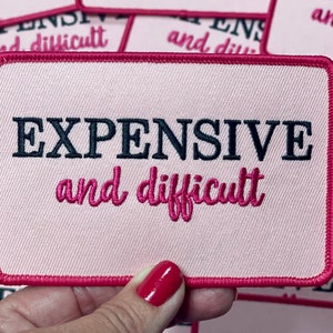 Op de afbeelding: Roze rechthoekige patch met de woorden "EXPENSIVE" in zwart en "and difficult" in roze script. De patch heeft een roze rand en wordt door een hand vastgehouden. Andere vergelijkbare patches staan op de achtergrond.