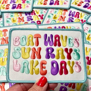 Puede incluir: Un parche blanco bordado con el texto "Boat Waves Sun Rays Lake Days" en letras de colores con forma de burbuja. El parche tiene un borde azul claro.