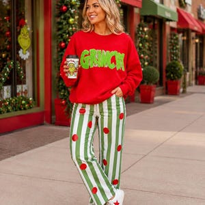 May include: A bright red long-sleeve sweatshirt with the word GRINCH in fuzzy green letters across the chest, paired with white and green vertical striped pants featuring red polka dots and white sneakers with a red star detail on the side.