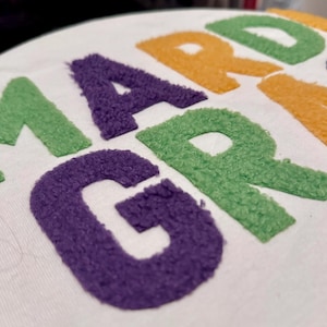 May include: Close-up of white fabric with the words "MARDI GRAS" embroidered in textured, colourful letters. The letters are in green, purple, and gold, evoking a festive and celebratory theme.
