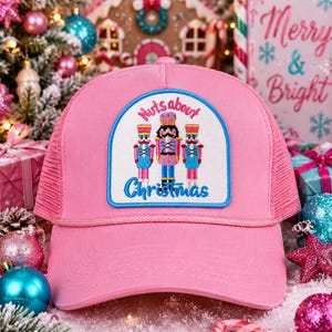 May include: A bright pink trucker hat featuring a white patch with a blue border. The patch displays three embroidered nutcracker figures in pink and blue, with the text "Nuts about Christmas" stitched in pink and blue thread above and below.
