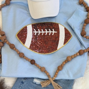 May include: Light blue sweatshirt with a sequined football design. The football is brown with white stripes and stitching. A white baseball cap rests above the football. Wooden bead garland and denim jeans complete the casual look.