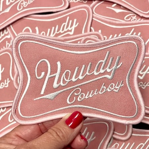 Może przedstawiać: Różowa łatka z tkaniny z białym haftem, na której widnieje napis "Howdy Cowboy" w retro stylu czcionki.