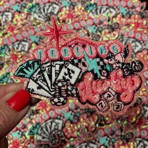 May include: A pink, glittery patch with the words "Feeling Lucky" in a stylized font. The patch features playing cards, dice, and a starburst design. The background is filled with similar patches.