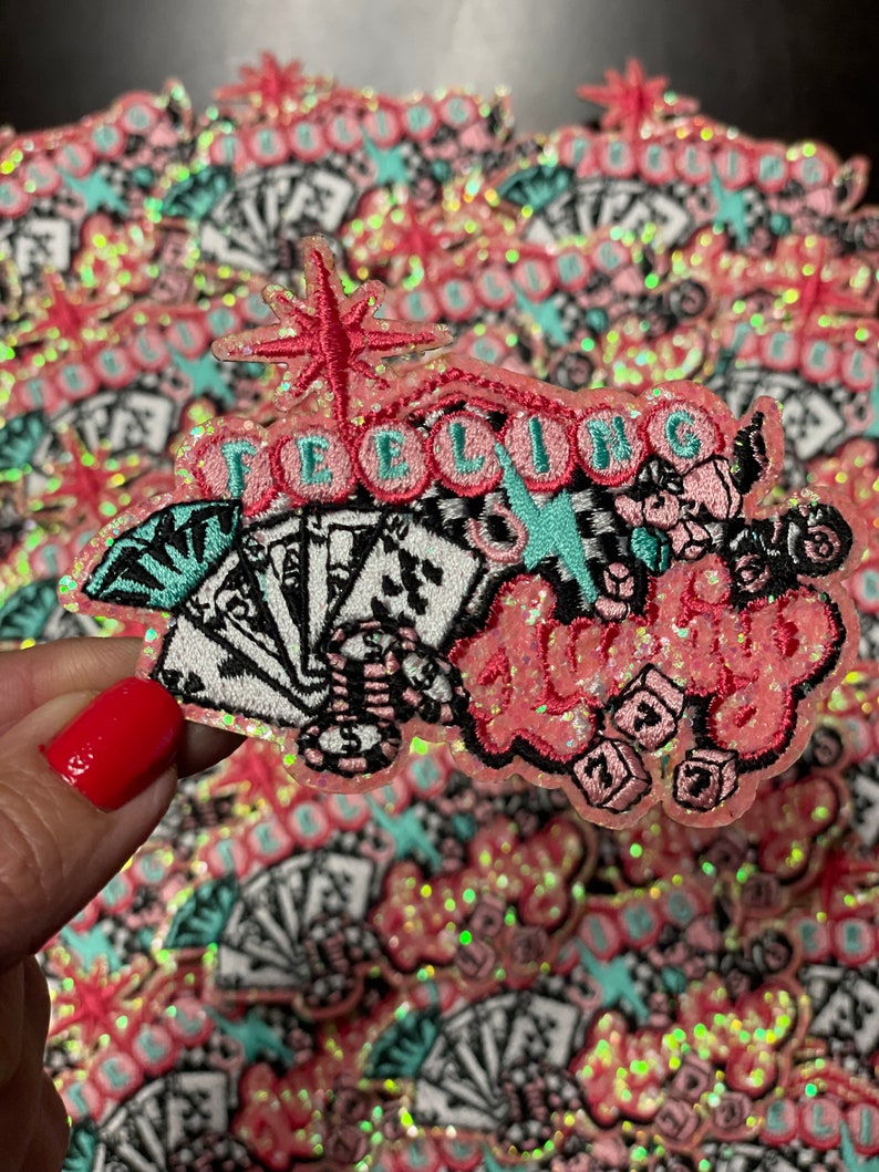 May include: A pink, glittery patch with the words "Feeling Lucky" in a stylized font. The patch features playing cards, dice, and a starburst design. The background is filled with similar patches.