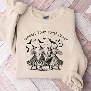 May include: A beige crewneck sweatshirt featuring a black graphic print of four figures wearing witch hats and dresses holding hands in a circle. Above the figures, the text reads Support Your Local Coven, with flying bats surrounding the scene.