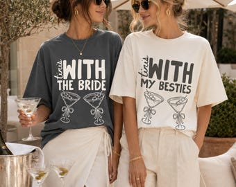 May include: Two short-sleeve crew neck t-shirts, one charcoal gray and one cream, featuring black text and line art of two martini glasses. The charcoal shirt reads "WITH THE BRIDE" and the cream shirt reads "WITH MY BESTIES".