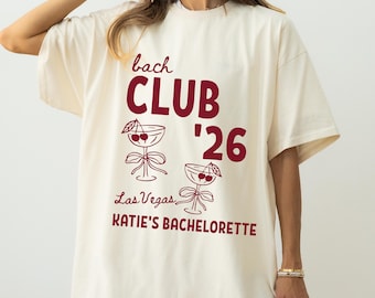 May include: An off-white short-sleeve t-shirt featuring dark red text that reads bach CLUB '26, Las Vegas, and KATIE'S BACHELORETTE. The design includes two line-art illustrations of stemmed cocktail glasses garnished with cherries and bows.