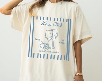May include: An off-white short-sleeve t-shirt featuring a blue graphic print of two wine glasses, grapes, and the text Wine Club, 2026, Las Vegas, and Taylor's Bachelorette, all framed by vertical blue stripes on the sides of the design.