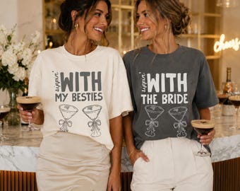 May include: Two crew neck t-shirts, one cream and one charcoal gray, featuring the text 'Sippin' WITH MY BESTIES' and 'Sippin' WITH THE BRIDE' respectively, accompanied by line art illustrations of two martini glasses with bows on the stems.