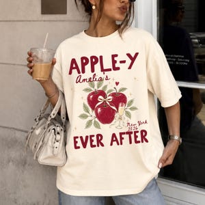 May include: A cream-colored short-sleeve t-shirt featuring a graphic of three red apples tied with a white ribbon, a champagne flute, and the text "APPLE-Y Amelia's EVER AFTER New York 2026" printed in dark red lettering with small gold star accents.