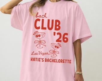 May include: A light pink short-sleeve t-shirt featuring red text that reads bach CLUB '26, Las Vegas, and KATIE'S BACHELORETTE. The design includes two line-art illustrations of stemmed cocktail glasses adorned with cherries and ribbons.