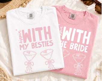 May include: A white t-shirt with pink text reading Sippin With My Besties and two martini glass graphics, and a pink t-shirt with white text reading Sippin With The Bride and a martini glass graphic, both featuring Comfort Colors neck labels.