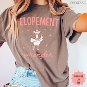 May include: Espresso-coloured t-shirt with the words "ELOPEMENT" and "wrangler" in pink script. A cartoon goose wearing a cowboy hat and boots holds a lasso. Pink star accents are scattered around the goose. The words "THERAPIST LIFE TEE" are in a pink starburst.