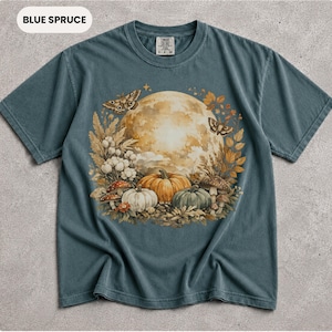 Puede incluir: Una camiseta de manga corta de color azul grisáceo con un estampado gráfico de una gran luna llena rodeada de follaje otoñal, polillas y calabazas. El diseño incluye calabazas naranjas, blancas y verdes con varias setas y hojas. La camiseta tiene cuello redondo y una etiqueta en el interior del cuello.