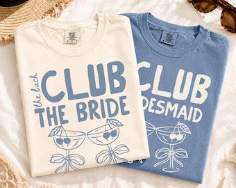 May include: Two short-sleeve crew neck t-shirts. The cream shirt features blue text reading the bach CLUB THE BRIDE with a graphic of two stemmed glasses with cherries. The blue shirt features white text reading CLUB BRIDESMAID with a glass graphic.