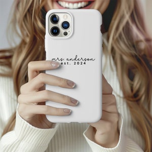 May include: White phone case featuring the text "mrs. Anderson est. 2024" in a script font. The phone case has a camera cutout and is held by a person wearing a white jumper.