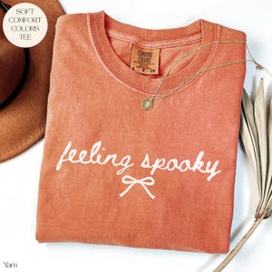 May include: A coral-coloured t-shirt with the words "feeling spooky" in white script and a bow graphic. A gold necklace with a pendant rests on the shirt. The shirt has a Comfort Colors tag.