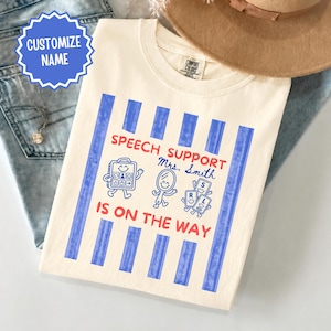 May include: A cream-colored t-shirt featuring vertical blue stripes and red text that reads "SPEECH SUPPORT IS ON THE WAY" along with "Mrs. Smith". The graphic includes illustrations of a speech device, a handheld mirror, and letter tiles.
