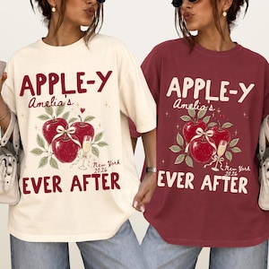 May include: A cream-colored t-shirt and a maroon t-shirt, both featuring the text APPLE-Y AMELIA'S EVER AFTER NEW YORK 2026. The graphic includes three red apples with green leaves, a white ribbon bow, a champagne flute, and small star accents.