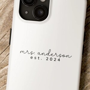 May include: White phone case with a black camera module. The case features the text "mrs. anderson est. 2024" in a script font. The top of the image has the text "BRIDE PHONE CASE CUSTOMIZABLE!"