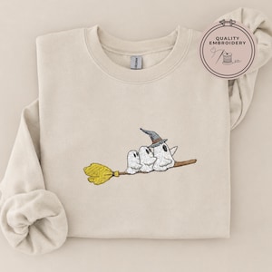 May include: A beige crewneck sweatshirt featuring an embroidered design of three white ghosts riding a brown broomstick. The largest ghost wears a grey witch hat. A circular badge in the top right corner contains the text Quality Embroidery.