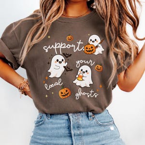 May include: A dark brown t-shirt featuring a graphic print with the text "support your local ghosts" in white script. The design includes three white cartoon ghosts, orange jack-o'-lantern pumpkins, small black bats, and yellow stars.