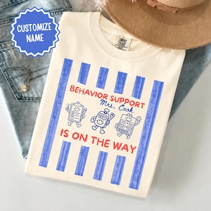 Op de afbeelding: Een crèmekleurig t-shirt met verticale blauwe strepen en rode tekst die BEHAVIOR SUPPORT IS ON THE WAY luidt. Het ontwerp bevat de gepersonaliseerde naam Mrs. Cook en drie stripfiguren: een klembord, een timer en een tokenbord.