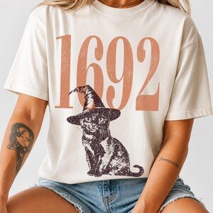 May include: A cream-colored short-sleeve t-shirt featuring the large, distressed-style terracotta-colored text 1692 printed across the chest. Below the numbers is a detailed black graphic illustration of a cat wearing a pointed witch hat.
