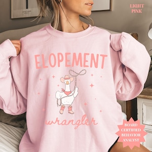 Op de afbeelding: Lichtroze sweatshirt met het woord "ELOPEMENT" in lichtroze blokletters. Daaronder een cartoon gans met cowboyhoed, laarzen en een halsdoek, die een lasso vasthoudt. Het woord "wrangler" staat eronder in cursief.