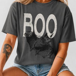 May include: A charcoal gray short-sleeve t-shirt featuring the word BOO in large, distressed white capital letters. Below the text is a detailed black graphic illustration of a bat with its wings spread, centered on the front of the shirt.