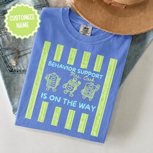 Könnte beinhalten: Ein kornblumenblaues T-Shirt mit vertikalen hellgrünen Streifen und einem hellblauen Grafikdruck. Der Text lautet BEHAVIOR SUPPORT Mrs. Cook IS ON THE WAY, ergänzt durch Illustrationen eines Klemmbretts, eines Timers und einer Token-Tafel.