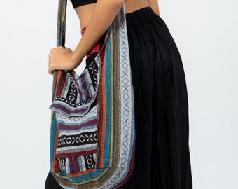 May include: A colorful shoulder bag with a patterned design in shades of blue, red, and brown. The bag has a long strap with a repeating geometric pattern. The person in the image is wearing a black top and skirt.