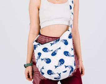 May include: White shoulder bag with a blue whale pattern. The bag has a curved shape and a long strap. The whale design features blue whales with striped bodies and yellow polka dots on a white background. The bag is worn over the shoulder.