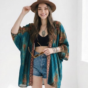 May include: A woman wearing a brown hat, a teal kimono with a patterned design, denim shorts, and brown cowboy boots. She is also wearing a black top and holding sunglasses.