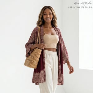May include: A beige crop top, white wide-leg trousers, and a burgundy kimono with a white mandala print. A woven straw tote bag is carried on the shoulder, and brown strappy sandals are worn. Text reads: Home 64 Studio MODEL TALL 175 CM/ 5.9 FEET.
