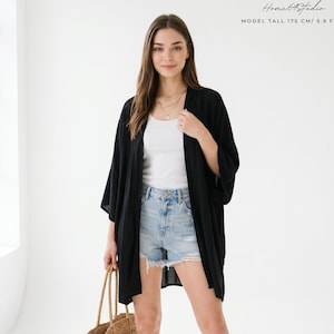 May include: A black open-front kimono cardigan worn over a white tank top and light blue distressed denim shorts. The outfit includes tan suede ankle boots and a woven straw tote bag. Text in the image reads: Home 64 Studio MODEL TALL 175 CM/ 5.9 FEET.