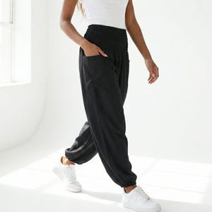 Puede incluir: Una mujer con una camiseta sin mangas blanca y pantalones harén negros con bolsillos. También lleva zapatillas blancas. El conjunto es informal y cómodo, adecuado para el día a día.